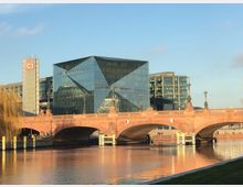 Moderne Glasfassade eines Bürogebäudes mit prismatischen Formen, flankiert von einem Gebäude mit DB-Logo, im Hintergrund. Im Vordergrund eine historische Steinbrücke mit dekorativen elementaren Statuen, die sich über einen ruhigen Fluss spannt.