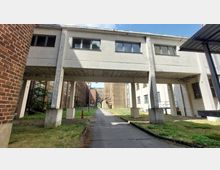 Industriekomplex mit gemauerten und verputzten Gebäuden, verbunden durch einen überdachten Übergang aus Beton auf Pfeilern. Im Vordergrund eine asphaltierte Straße und Grünflächen, die zu weiteren Backstein- und Betonbauten führen.