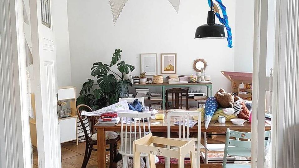 A cluttered dining area with a wooden table surrounded by mismatched chairs, a children's step stool in the foreground, and a rug covering wooden parquet flooring. The room is decorated with white triangular bunting, potted plants, framed wall art, and a black hanging light fixture.
