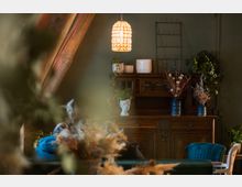 Wohnraum mit rustikalem Holzschrank im Hintergrund, darauf Dekorationen wie Keramikvasen, trockene Blumensträuße und Pflanzen. Die Szene ist in warmem Licht beleuchtet, mit Holzbalken und einer hängenden Lampe im Vordergrund.