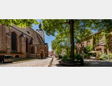 Gepflasterte Gasse mit historischer Backsteinkirche links, gekennzeichnet durch hohe gotische Fenster und ein Kirchendach mit Ziegeln; rechts ein von Bäumen beschatteter Hof mit einem begrünten Backsteingebäude, Tischen und Sonnenschirmen.