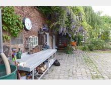 Gepflasterter Hinterhof mit einer Backsteinmauer, an der eine Wanduhr und Efeu sowie ein blühender Flieder hängen. Ein Arbeitstisch mit Blumentöpfen und Gartenutensilien steht an der Wand, während im Hintergrund eine grüne Gartenlandschaft mit Bäumen und Sträuchern sichtbar ist.