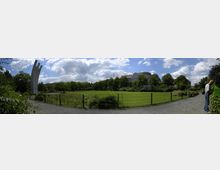 Panorama eines Parks mit einer großen, offenen Rasenfläche, umgeben von Bäumen und Büschen. Links steht ein modernes Denkmal mit drei vertikalen Elementen, während im Hintergrund ein historisch wirkendes Gebäude sichtbar ist; ein Spazierweg verläuft entlang eines Zauns im Vordergrund.