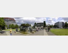 Panoramablick auf einen Platz in einer Stadt mit einem Brunnen im Zentrum, umgeben von Skulpturen. Im Hintergrund sind grüne Rasenflächen, Bäume sowie mehrere Gebäude, darunter ein Kaufhaus mit der Aufschrift „KaDeWe“.