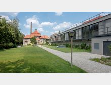 Grüne Außenanlage mit einem gepflasterten Weg, der zu einem historischen Gebäude mit rotem Ziegeldach und hohem Schornstein führt. Rechts davon ein moderner, langgezogener Bau mit Glasfronten und bepflanztem Umfeld.