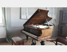 Ein Konzertflügel mit geöffneter Klavierhaube aus Holz steht in einem schlichten Raum mit Holzboden. Im Hintergrund sind eine helle Wand mit einem Bild, ein Fenster mit Vorhang sowie ein Holzschrank zu sehen.