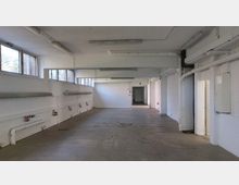 Leerer, rechteckiger Raum mit weiß gestrichenen Wänden, sichtbaren Rohrleitungen und mehreren Steckdosen entlang der Wände. Der Boden besteht aus Beton, und nahe der Decke befinden sich mehrere Fenster, die Tageslicht hereinlassen.