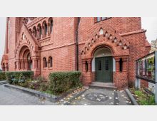 Backsteinfassade einer historischen Kirche im neugotischen Stil mit rundbogigen Fenstern, verziertem Portal und grüner zweiflügeliger Metalltür. Im Vordergrund befindet sich ein gepflasterter Gehweg mit einem kleinen bepflanzten Bereich und Büschen, rechts neben dem Eingang sind Glas-Schaukästen mit Veranstaltungsplakaten sichtbar.