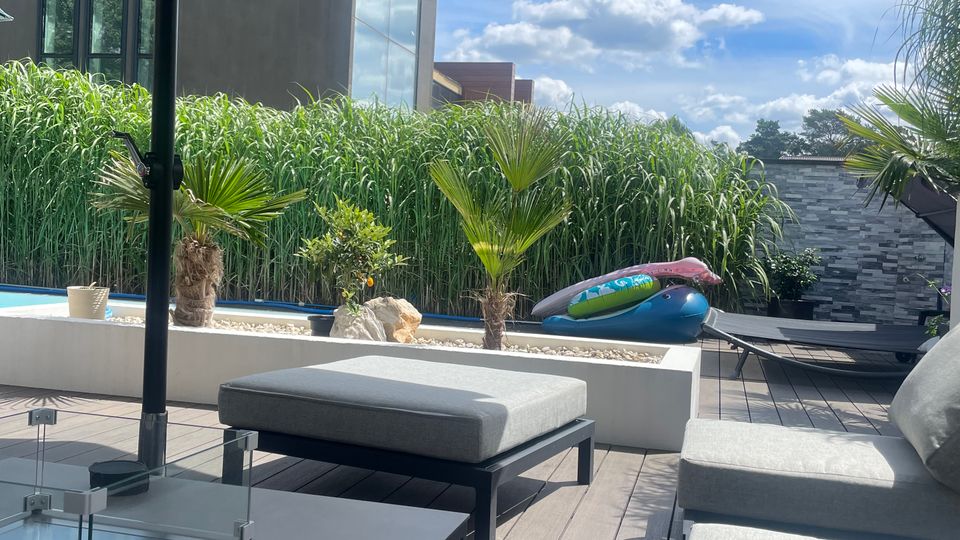 Moderne Dachterrasse mit Holzboden, mehreren grauen Polstermöbeln und einem großen Sonnenschirm. Im Hintergrund befinden sich Pflanzen, ein kleiner Pool und ein Haus mit großen Glasfenstern.