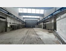 Große, leere Industriehalle mit hohen Decken, freiliegenden Stahlträgern und einer Glasdachkonstruktion für Tageslichteinfall. Sichtbare Schienen verlaufen entlang des Betonbodens, und die Wände bestehen aus unverputztem Mauerwerk.