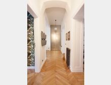 Flur in einer Wohnung mit Holzfußboden im Fischgrätmuster, weißen Wänden und einer geschwungenen Deckenarchitektur. Am Ende des Flurs befindet sich eine Metalltür, und an den Wänden hängen Bilder sowie ein schwarzer Heizkörper. Links ist eine Tapete mit tropischem Muster zu sehen.