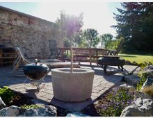 Steinpatio mit rustikaler Steinmauer im Hintergrund, ausgestattet mit Gartenmöbeln, darunter ein Holztisch mit Stühlen, ein Grill und ein Feuerkorb. Im Vordergrund sind ein runder Pflanztopf und blühende Blumenbeete zu sehen, umgeben von einer natürlichen Gartenlandschaft mit Bäumen und Wiese.