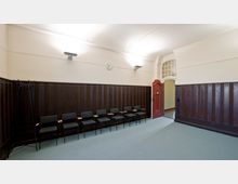 Ein schlicht eingerichteter Raum mit dunkler Holzvertäfelung an den Wänden, mehreren gepolsterten Stühlen in einer Reihe, einer roten Tür mit gewölbtem Türrahmen und einer Wanduhr über der Vertäfelung. Der Boden ist mit hellgrünem Teppich ausgelegt, und die Beleuchtung erfolgt durch Wandlampen.