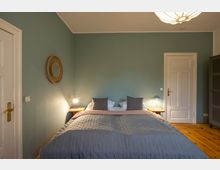 Schlafzimmer mit einem Doppelbett vor einer hellgrünen Wand, flankiert von zwei Nachttischen mit modernen Lampen. Holzfußboden, weiße Türen und ein rundes Wanddekor aus Rattan ergänzen die schlichte Einrichtung.