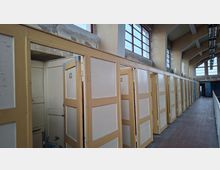 Ein alter Innenraum mit einer Reihe gelblicher Holzkabinen mit nummerierten Türen, darunter teilweise offene Türen. Die Wände sind verputzt und stellenweise abgenutzt, und oben verläuft eine Reihe von Fenstern mit roten Rahmen.