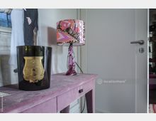 Ein Innenraum mit einem lila Samttisch, auf dem eine schwarze Duftkerze mit goldenem Etikett und eine Lampe mit gemustertem, farbenfrohem Lampenschirm stehen. Im Hintergrund eine weiße Tür und ein teilweise sichtbares Foto.