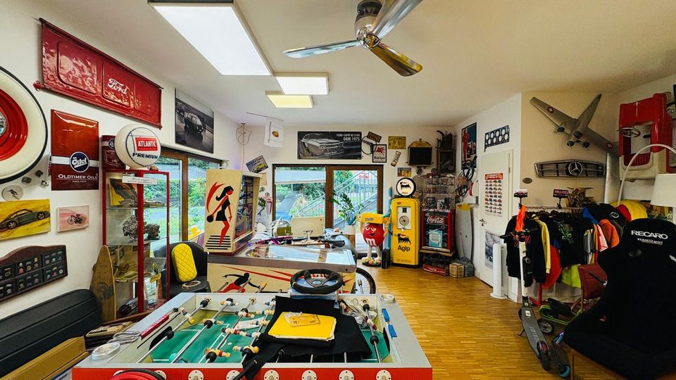 Innenraum eines hobbyartigen Werkstatt- und Sammlerraums mit Holzfußboden, Deckenventilator und großen Fenstern; an den Wänden hängen Vintage-Auto- und Tankstellen-Schilder, Teile und Dekoration. Im Vordergrund steht ein Tischkicker, daneben Vitrinen und Regale mit Gegenständen sowie ein Kleiderständer und diverse Fahrzeugteile.