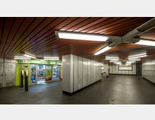Unterirdischer U-Bahn-Zugang mit beleuchteter Decke aus braunen Holzpaneelen, hellem Metall an den Wänden und einem kleinen Kiosk mit einer grünen Fassade auf der linken Seite. Im Hintergrund befinden sich Wegweiser und Fahrpläne.