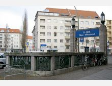 Eingang zur U-Bahn-Station „Neu-Westend“ in Berlin, mit einem blauen Schild über dem Zugang. Der Bereich ist von Geländern und zwei steinernen Säulen mit Lampen eingefasst, im Hintergrund befinden sich mehrstöckige Wohngebäude mit roten Ziegeldächern.