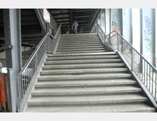 Treppe unter einer Brückenkonstruktion mit Metallgeländern an beiden Seiten. Die Umgebung zeigt Stahlträger und Säulen, teilweise mit Graffiti bemalt, sowie eine Person, die die Treppe hinaufgeht.