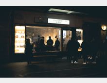 Straßenansicht eines kleinen Kinos oder Cafés bei Nacht. Mehrere Menschen stehen vor dem beleuchteten Schaufenster, das Regale mit Büchern oder Plakaten zeigt, während im Inneren weitere Personen an einem Tisch sitzen. Oberhalb des Eingangs ist ein Schild mit der Aufschrift „Fava Connection“ angebracht.