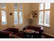 Wohnzimmer mit klassischen roten Ledersesseln, rundem Holztisch und Stehlampe. Helle Wände mit großen Fenstern, durch die Bäume im Außenbereich sichtbar sind, sowie ein Heizkörper unter den Fenstern und ein Gemälde an der Wand.