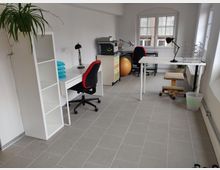 Ein heller Büroraum mit grauem Fliesenboden, zwei Schreibtischen mit Schreibtischlampen und Bürostühlen, einem weißen Regal, einem gelben Rollcontainer und einem Gymnastikball. Große Fenster lassen Tageslicht herein, und eine Topfpflanze steht auf dem Regal.