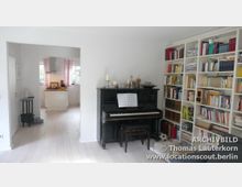 Wohnzimmer mit einem schwarzen Klavier an der Wand, verziert mit Kerzen und Dekorationen. Daneben steht ein großes weißes Bücherregal, gefüllt mit Büchern und anderen Gegenständen. Im Hintergrund ist eine offene Tür, die in eine helle Küche mit rosafarbenen Vorhängen führt.