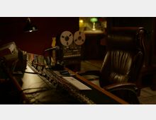 Ein Tonstudio mit einem Mischpult im Vordergrund, einem Mikrofon und einem Computer-Tastatur-Maus-Setup. Im Hintergrund sind ein analoger Bandrekorder und eine grüne Schreibtischlampe sichtbar, die Szene ist in warmem, gedämpftem Licht gehalten.