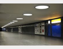 Unterführung mit rechteckigem Grundriss, beleuchtet durch runde Deckenlampen. Wände sind mit dunklen Fliesen verkleidet und zeigen Plakate; ein gelbes Schild markiert den Ausgang "Zimmermannstraße".