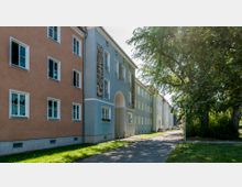 Wohnhäuser in einer Reihenbebauung mit verschiedenen pastellfarbenen Fassaden, großen Fenstern und teils dekorativen Wanddetails; im Vordergrund ein Gehweg, angrenzend eine Grünfläche mit Bäumen.