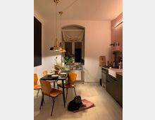 Ein gemütlicher Küchen- und Essbereich mit hellem Holzboden, einem schwarzen Esstisch, umgeben von vier Holzstühlen. Im Hintergrund ein Fenster mit weißem Stoffvorhang, umgeben von Pflanzen; rechts eine moderne Küchenzeile mit Spüle, Herd und hölzerner Arbeitsfläche. Ein kleiner Hund liegt auf einem rosa Kissen auf dem Boden.