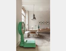 Ein industriell gestalteter Raum mit einer groben Betondecke, großen Fenstern auf der linken Seite und unverputzten Wänden. Dominierend sind eine grüne Retro-Bodenwaage im Vordergrund, ein rustikaler Tisch aus Holz und Betonklötzen sowie eine Hängelampe aus Metall über dem Tisch.