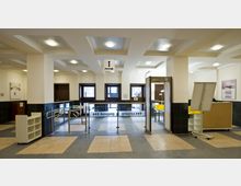 Eingangsbereich eines öffentlichen Gebäudes mit moderner Sicherheitskontrolle, bestehend aus Drehkreuzen, einem Metalldetektor und Hinweistafeln. Der Raum hat helle Wände, dunkel geflieste Sockel, eine Kassettendecke mit Leuchten und einen gefliesten Boden.