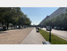 Breite Allee mit gepflastertem Gehweg in der Mitte, flankiert von Bäumen, Stadtgebäuden und einer Autostraße auf beiden Seiten; im Hintergrund ist ein Turm erkennbar. Auf den Gehwegen stehen Bänke, und vereinzelt sind Fußgänger, Autos und Fahrräder zu sehen.