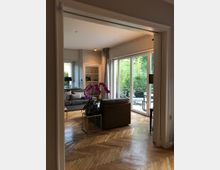 Wohnzimmer mit Holzparkettboden und großen Fenstern zum Garten. Im Raum stehen ein Sofa, ein Sessel und ein kleiner Tisch mit Orchideen, während der Hintergrund einen Sitzbereich im Freien zeigt.