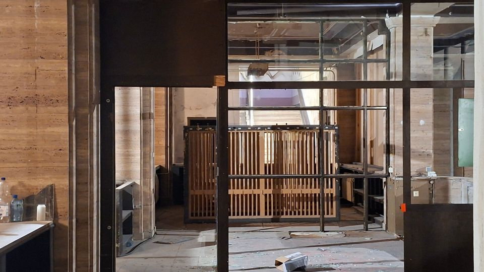 Innenraum eines im Umbau befindlichen Gebäudes mit Holzpaneelwänden und großen, schwarz gerahmten Glas- und Metalltrennwänden. Im Hintergrund stehen Holzgeländer und offene Deckeninstallationen, am Boden liegen Bau- und Verpackungsreste.