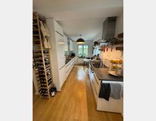 Moderne, schmale Küche mit Holzfußboden, weißen Hochglanzmöbeln und schwarzen Arbeitsplatten. Links ein Weinregal, rechts ein Herd mit Dunstabzugshaube; im Hintergrund ein großes Fenster mit Tisch und Pflanzen.