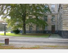 Steinernes Gebäude mit großen rechteckigen Fenstern und hellbrauner Fassade, umgeben von einem gepflasterten Gehweg und einer Grünfläche mit einem großen Baum. Im Vordergrund eine Straße, eine Laterne und ein Papierkorb.