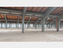 Großer, leerer Innenraum einer Industriehalle mit grauen Pfeilern und einem Dach aus Holz und Stahlträgern. Der Boden ist teilweise beschädigt, Fensterreihen an den Wänden lassen Tageslicht hinein.