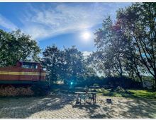 Wiese mit Bäumen und einer alten, rostigen Lokomotive auf einem Holzstapel; im Vordergrund stehen Tische und Stühle auf einem Kopfsteinpflaster, Sonnenschein und blauer Himmel bestimmen die Szene.