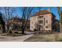 Wohngebäude im Stil der frühen Moderne mit beiger Fassade, rotbraunem Ziegeldach und mehreren Fenstern mit gelben Rahmen, umgeben von kahlen Bäumen und einer parkähnlichen Landschaft mit Gehwegen. Im Vordergrund führt ein geschwungener Weg durch eine offene Grünfläche.