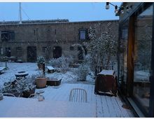 Schneebedeckter Innenhof mit Pflanzkübeln, Gartenmöbeln und Büschen vor einer historischen Backsteinfassade mit mehreren Fenstern und Türen. Im Vordergrund eine Holzterrasse mit einem Tisch und Stühlen, rechts ein modernes Gebäude mit Glasfassade.