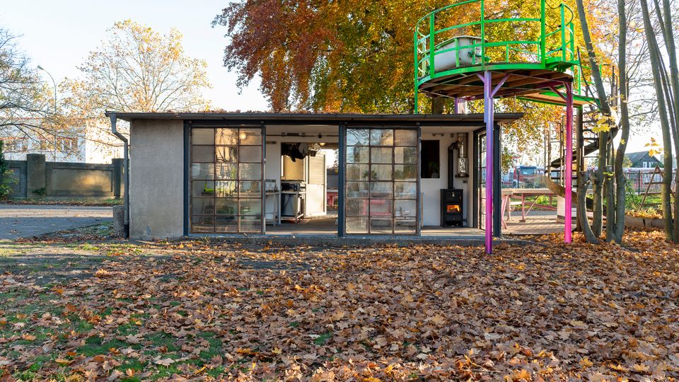 Kleines Gebäude mit großen Fenstern und einer offenen Front, umgeben von Herbstbäumen und gefallenen Blättern. Auf dem Dach befindet sich eine grün-gelbe Aussichtsplattform, die von pink-violetten Säulen gestützt wird.