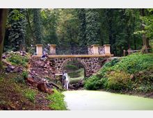 Steinbrücke mit Geländer aus Metall über einem kleinen, mit grünem Bewuchs bedeckten Bach in einem bewaldeten Park. Im Vordergrund ist ein Hang mit Erde und Felssteinen, während zwei Personen an der Brücke arbeiten.