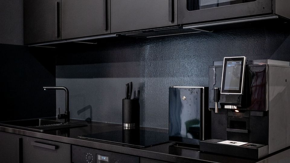Moderne, dunkelgraue Einbauküche mit matten Schrankfronten, schwarzer Arbeitsplatte und schlichter Rückwand. Sichtbar sind Spüle, Backofen und Mikrowelle sowie eine Kaffeemaschine auf der rechten Seite der Arbeitsfläche.