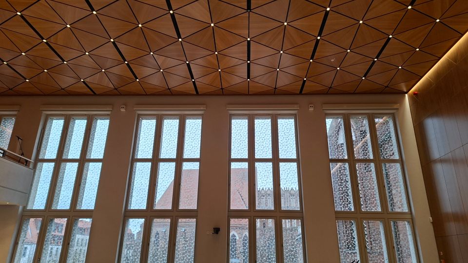 Innenraum eines modernen Saals mit hoher Decke aus dreieckigen Holzpaneelen und punktförmigen Leuchten. Große, hohe Fensterfronten mit gemustertem Glas lassen Tageslicht herein und geben den Blick auf Backsteinbauten und Kirchturmstrukturen draußen frei.