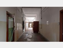 Ein langer, schlichter Flur in einem älteren Gebäude mit abgenutztem Boden, cremefarbenen Wänden und sichtbaren elektrischen Leitungen. Mehrere geöffnete Holztüren führen in angrenzende Räume, im Hintergrund ist Tageslicht durch Fenster sichtbar.