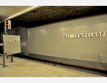 U-Bahn-Station mit gefliester Wand in hellem Grau, auf der das Schild "Alt-Tempelhof" zu sehen ist. Im Vordergrund steht ein rechteckiger Spiegel, der das Stationsschild spiegelverkehrt zeigt, neben Gleisen und einer Bahnsteigkante mit gelblichen Fliesen.