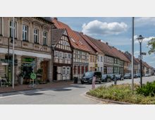 Straßenszene mit historischer Fachwerkhäuserreihe und einem Gebäude mit klassizistischer Fassade. Autos parken entlang der Straße, und im Vordergrund befindet sich ein Blumenladen mit Schildern und Dekorationen.